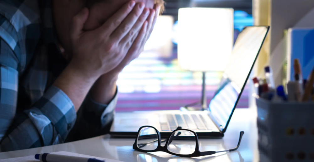 A man sat at an open laptop with his head in his hands, his glasses are sat on the desk 