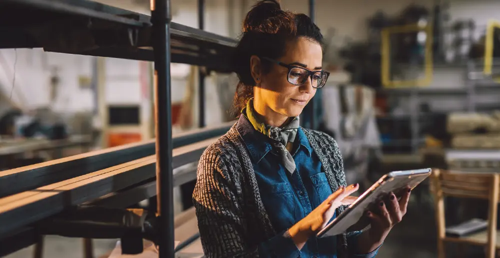 A woman standing in a workshop is holding an iPad, she is wearing a cardigan and her brown hair is up in a bun