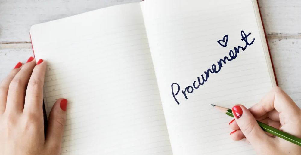 An open notebook with the word "Procurement" with a heart above the second "e" written in marker on the right hand page. A female hand with red painted nails holds the left page open and the other hand is holding a pencil on the right 