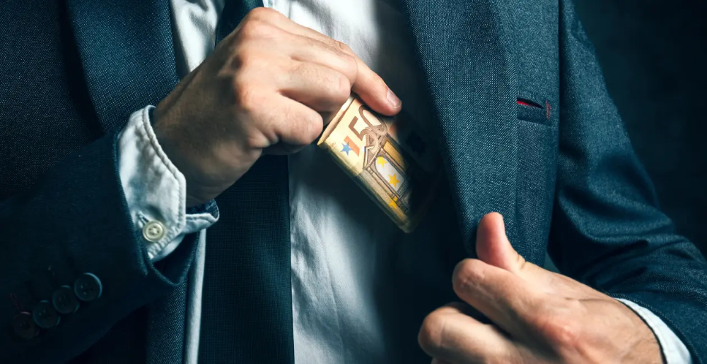 A close up of a mans chest wearing a shirt, tie and suit jacket, putting paper money into his inside jacket pocket