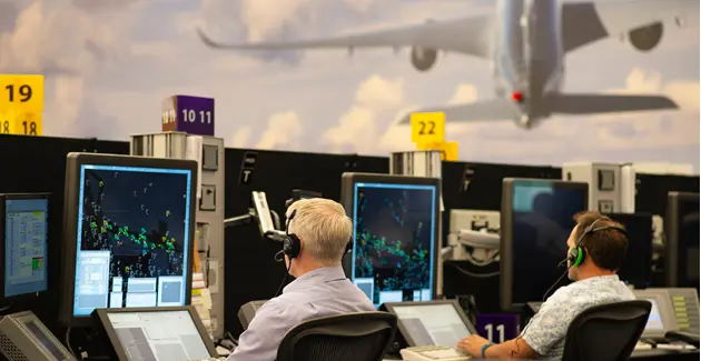 Air traffic control room with two men sat at control stations wearign headsets, a plane is taking off in the window above, away from the tower