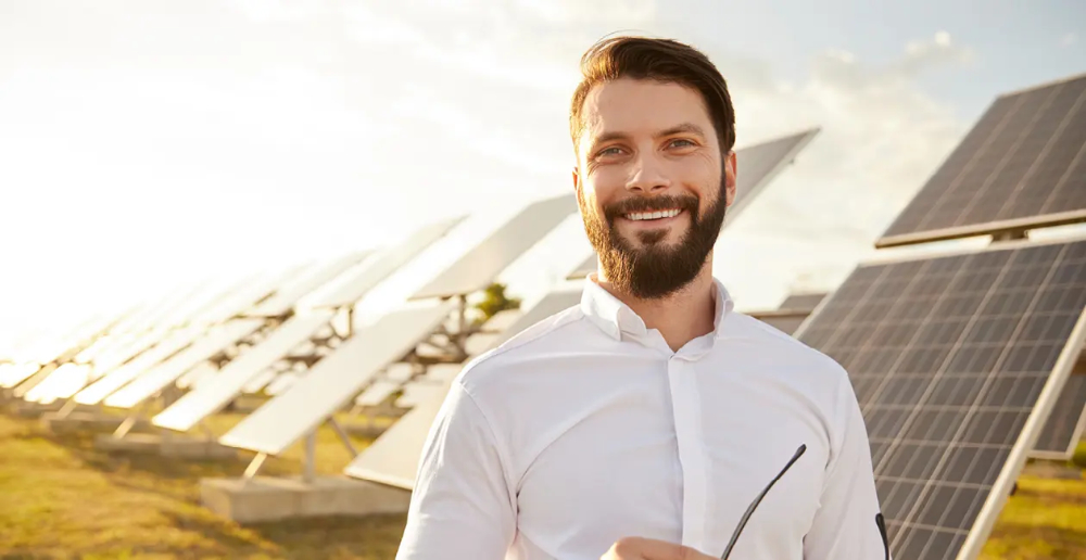 A man with a beard, wearing a white shirt and holding a pair of glasses, smiling and standing outside infront of solar panels