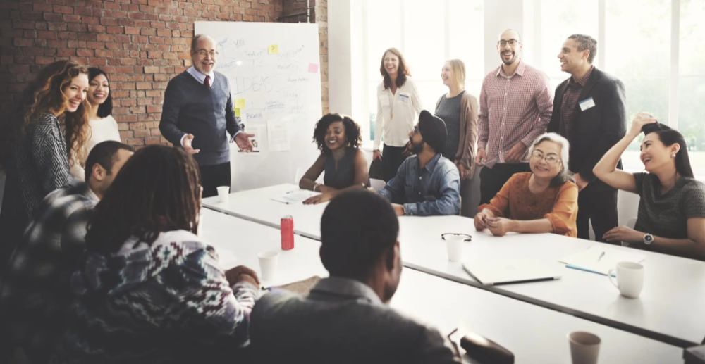 Lots of people sat and standing around a large conference table with one man standing at the head of the table with a whiteboard behind him. Everyone is smiling 