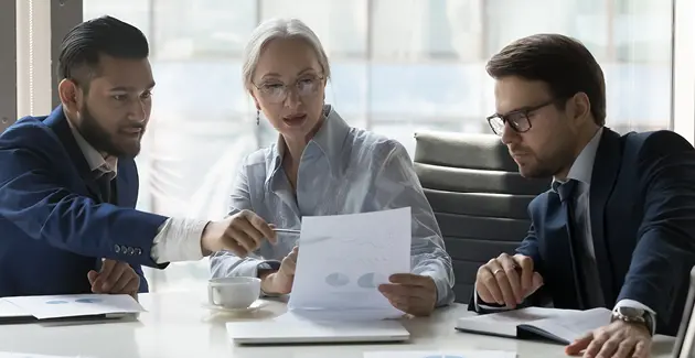 Three business people sat at a desk looking at a single piece of paper. One man is pointing with a pen to a part of it