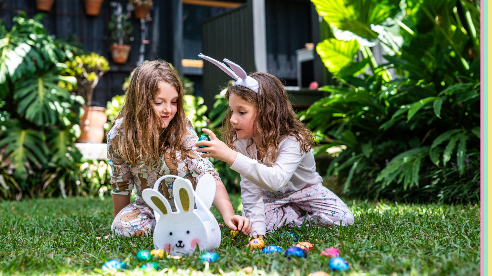 Two children in Easter themed pyjamas, one wearing bunny ears, are sitting on grass examining colorful Easter eggs near a bunny-shaped basket. Lush plants and hanging pots are in the background.