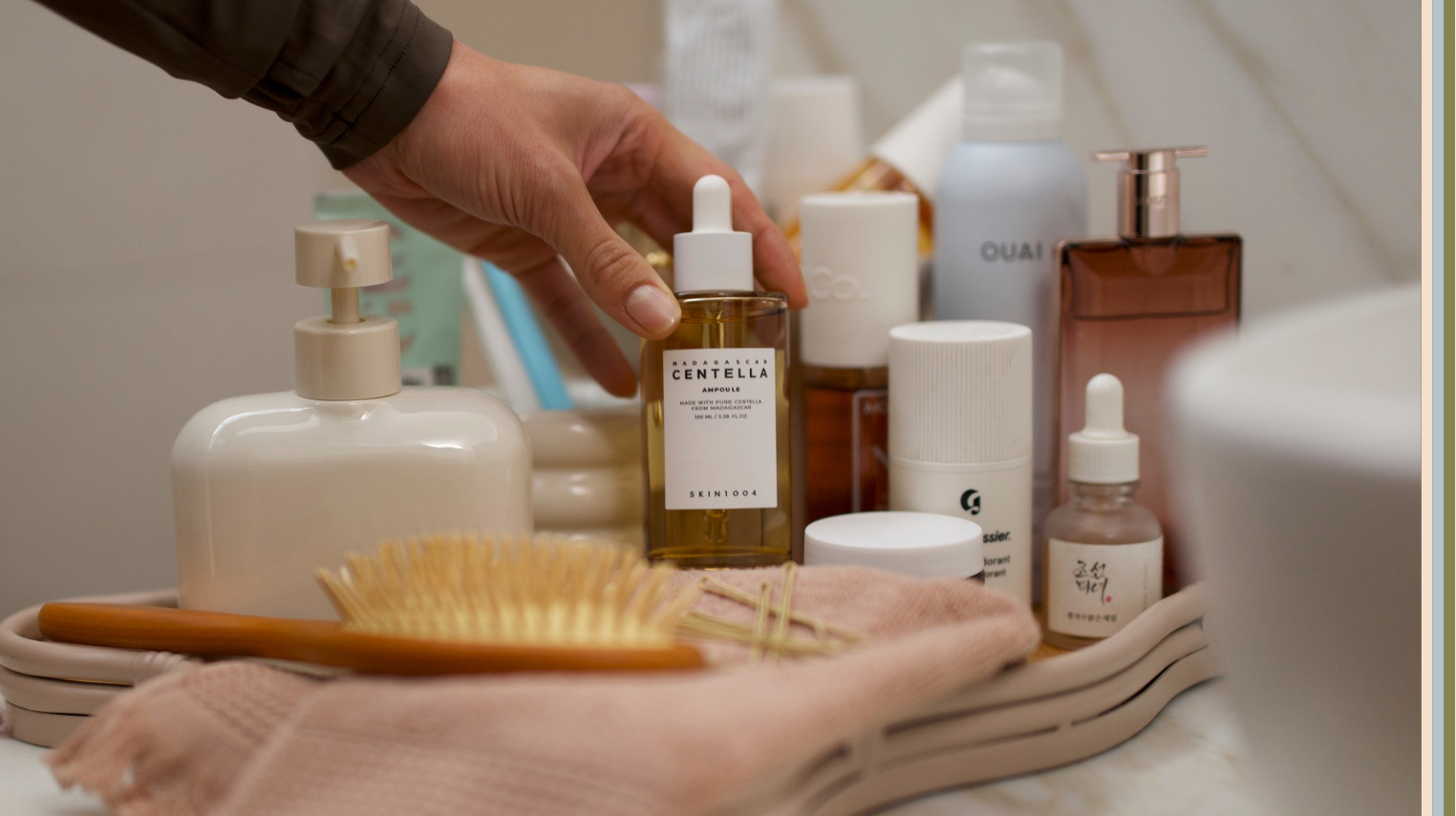 Hand arranging skincare products on bathroom tray with serum bottles, lotion dispenser, hairbrush, and beauty essentials for daily self-care routine