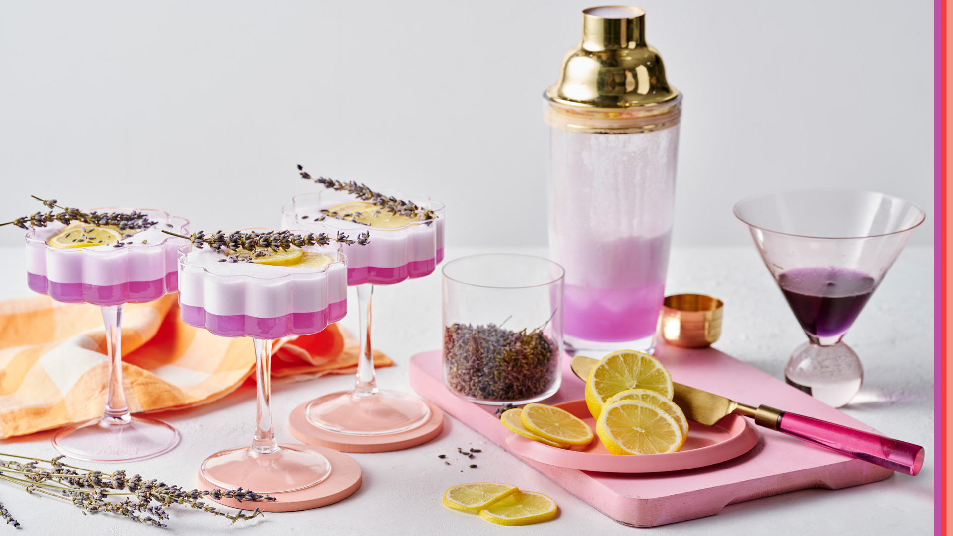 Three flower shaped cocktail glasses containing purple hued cocktail on white backdrop with cocktail shaker, lavender and lemon.