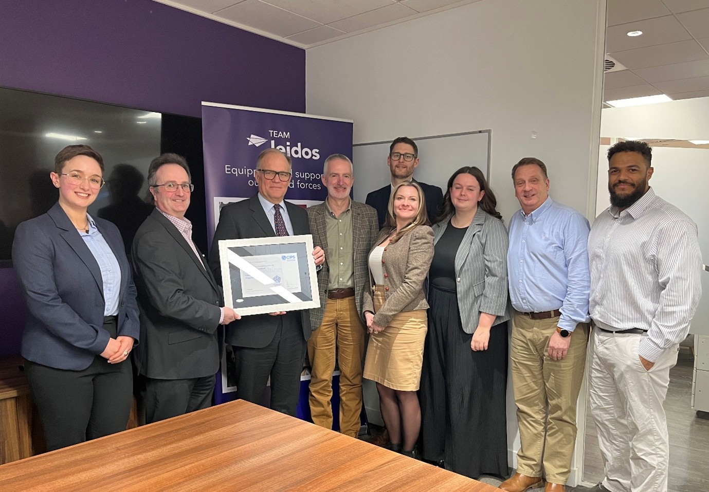 A group of nine colleagues stand together in a modern office space, smiling for a celebratory photo. A man in a dark suit and glasses on the left holds a white-framed CIPS recognition certificate. They are positioned in front of a purple Team Leidos branded pull-up banner. The group is dressed in a mix of business professional and business casual clothing. The room features a purple accent wall, a whiteboard, and a wooden meeting table.
