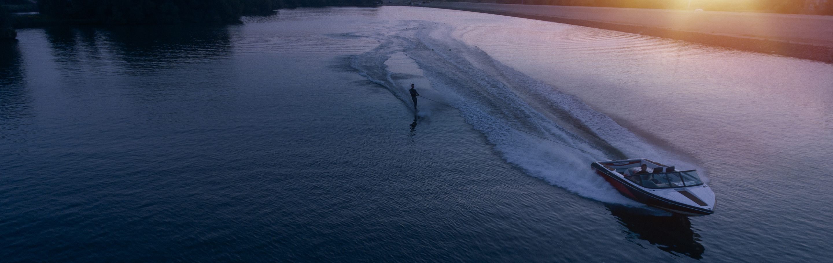 A speedboat with a water-skier sailing across a lake in the sunset