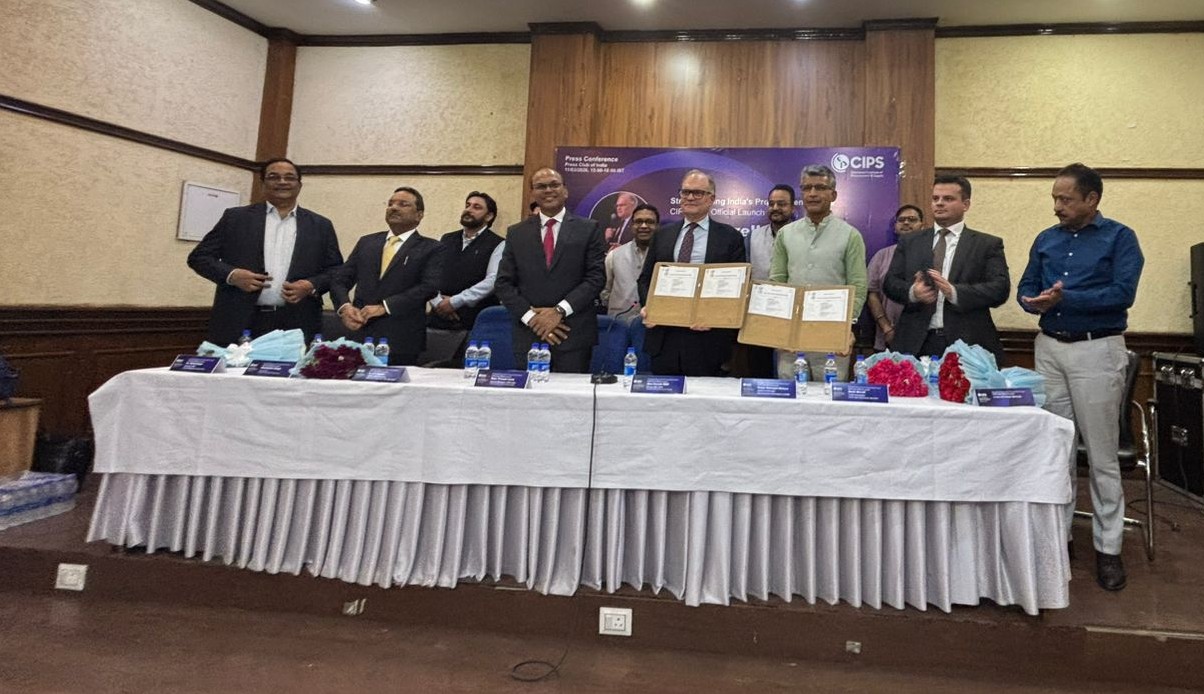 A group of men stand behind a long, white-skirted table in a wood-paneled room during a press conference. Two men in the center are holding up signed folders, likely a Memorandum of Understanding (MoU). A purple backdrop behind them features the CIPS (Chartered Institute of Procurement & Supply) logo and the text 