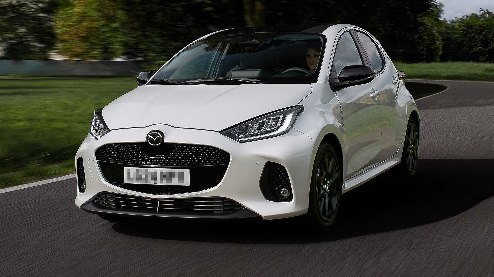 A white Mazda2 Hybrid compact hatchback is being driven on a curving road through a green landscape.