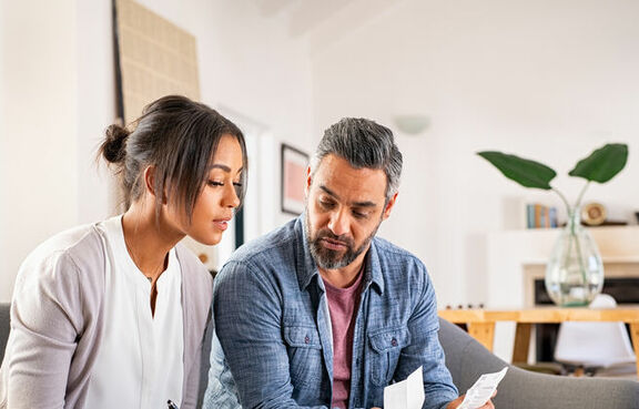 Middle aged couple sitting on a couch in front of a computer, looking at financial documents