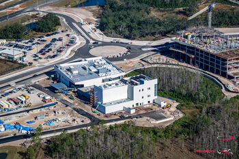 An aerial photo shows a construction site with a white building in the foreground surrounded by construction activities. The background includes a building framed with rebar and freshly paved roads.