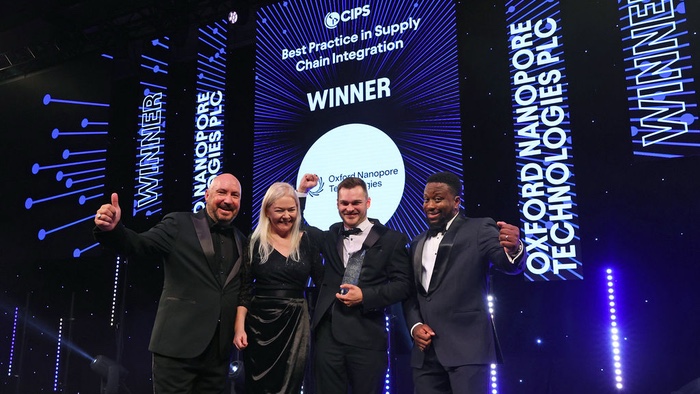 Image of two members of the Oxford Nanopore Technologies team on stage celebrating their award win holding the award. On either side are the host of the event (right) and a member of the CIPS team (left). The four people are standing on stage in front of blue graphics behind them on big screens saying the company's name, the award and 'Winner". 