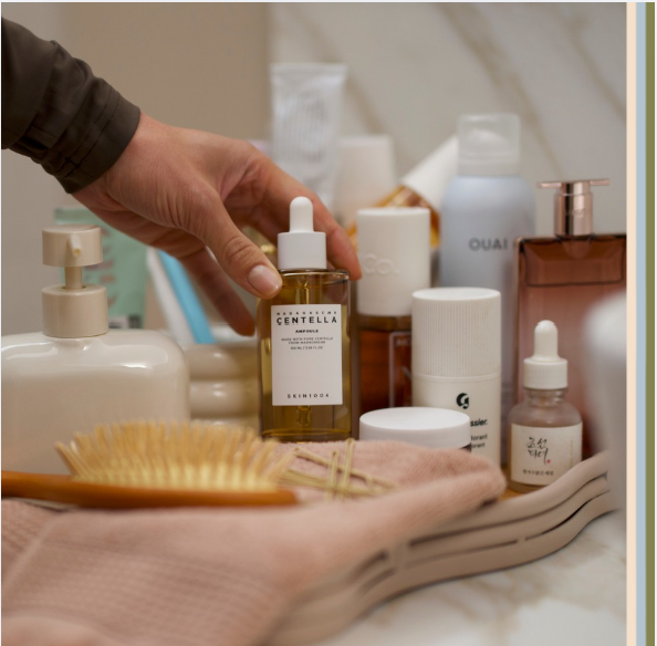 Hand arranging skincare products on bathroom tray with serum bottles, lotion dispenser, hairbrush, and beauty essentials for daily self-care routine