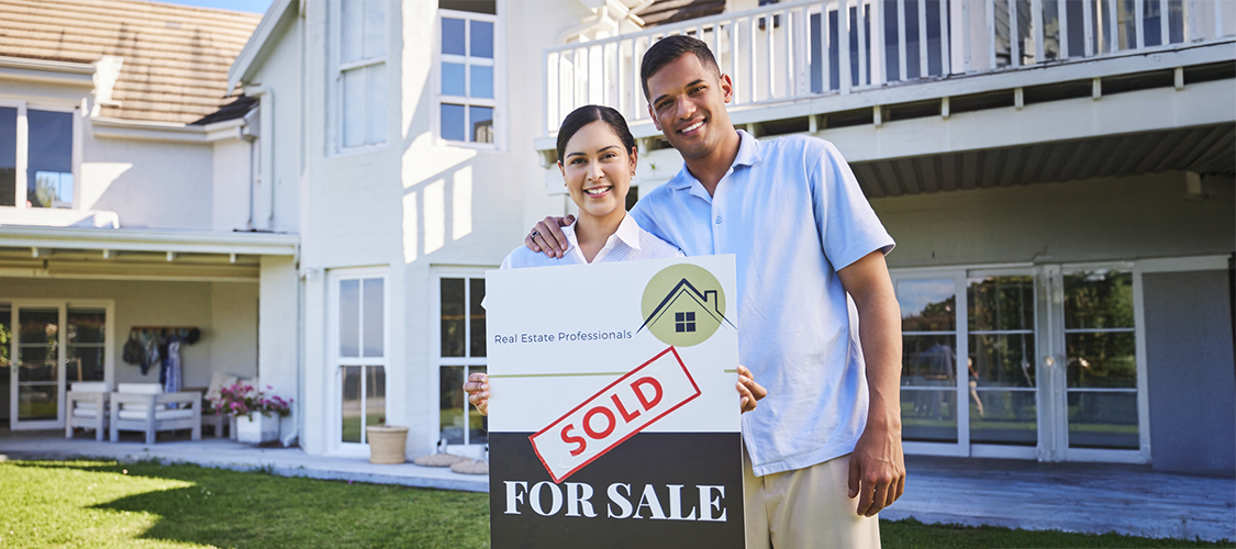 A couple standing outside of a home with a 'Sold' sticker on a 'For Sale' sign.