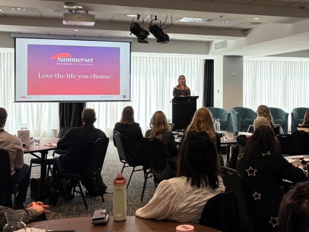 A speaker is standing behind a dark podium addressing an audience in a conference room. Behind her is a large screen displaying the text 'Summerset Retirement Villages. Love the life you choose' over a red and purple gradient background.