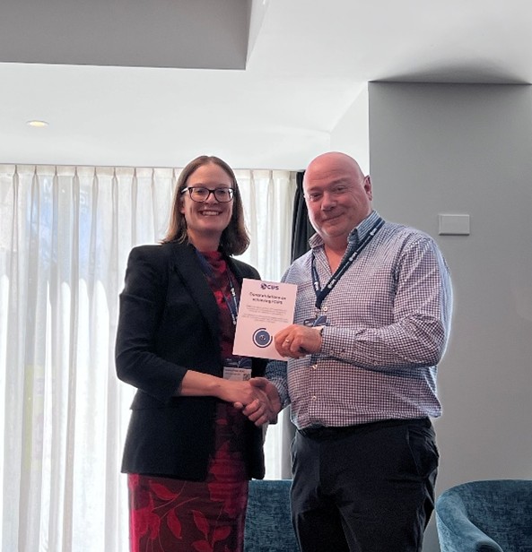 A woman in a red dress and dark blazer shaking hands with a bald man in a checked shirt. The man is holding a certificate from CIPS that says 'Congratulations on Renewing FCIPS.' They are standing indoors with a curtained background.