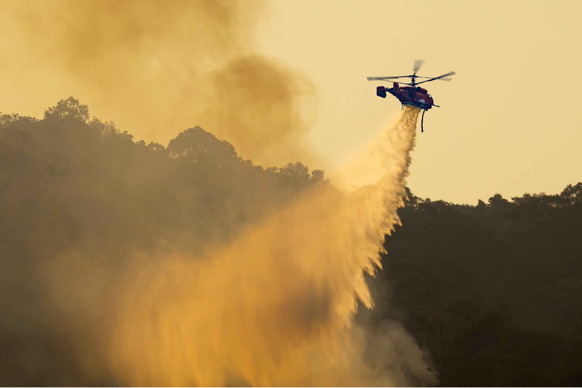 A firefighting helicopter dropping water over a smoky, tree-covered hillside during a wildfire under a hazy yellow sky.