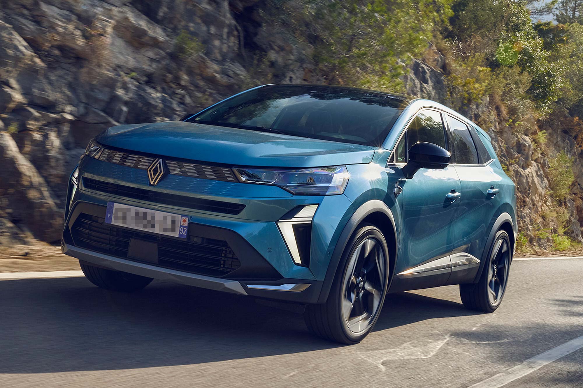 Blue Renault Symbioz SUV driving beside a rocky hillside, shown from the front left.