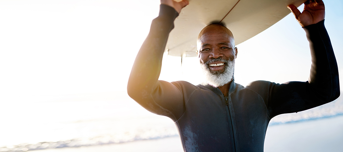 A senior Black man stands grinning on a beach in a wetsuit holding a surf board over his head