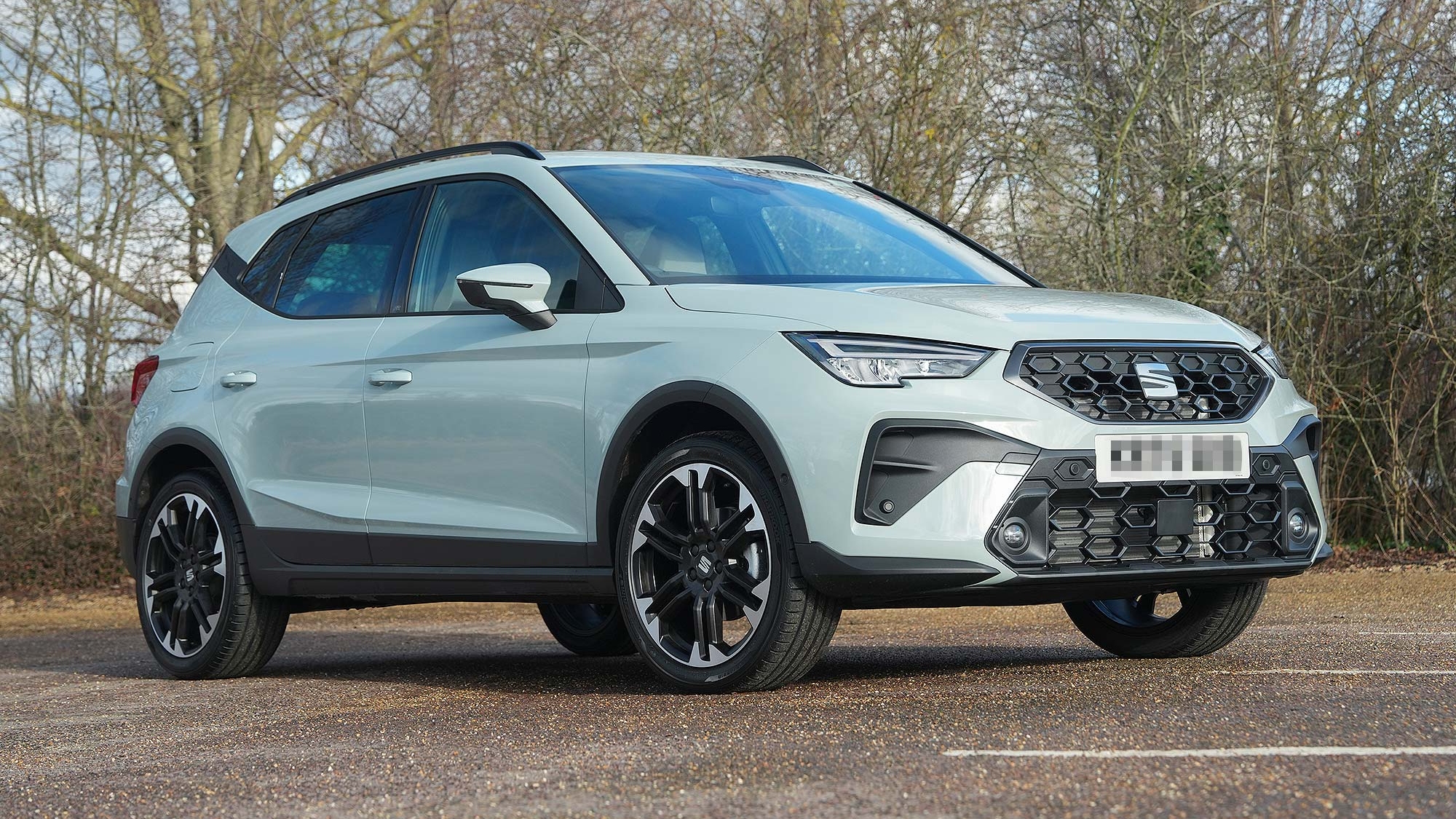 A light grey SEAT Arona parked on a paved area with trees behind, shown from the front three quarter angle, available on the Motability Scheme.