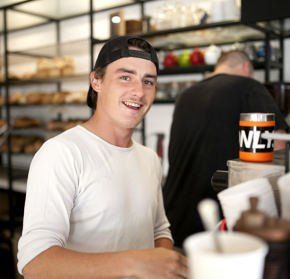 Smiling young barista in a white shirt and backwards cap behind the counter of a café.
