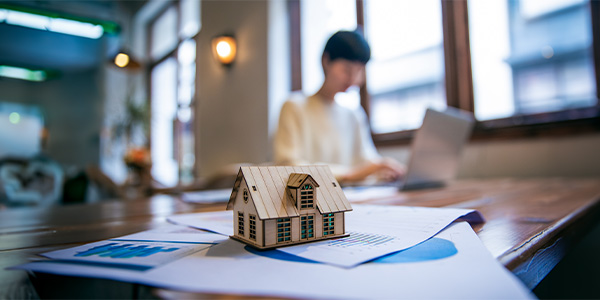 Someone is working on a laptop next to a stack of paperwork with a home on top of it.