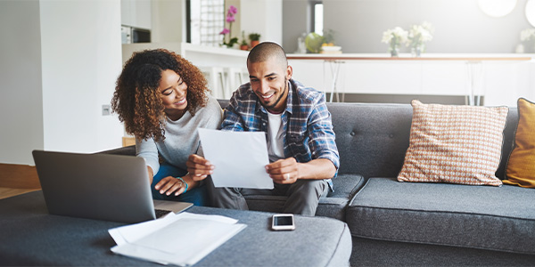 A Black man and Black woman sit together on a couch in a nice living room. They are smiling at a piece of paper the man is holding.  
