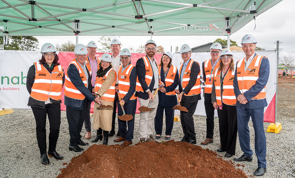 Group of people wearing safety vests and hard hats at a groundbreaking ceremony, smiling and holding shovels with soil in front of a construction site.