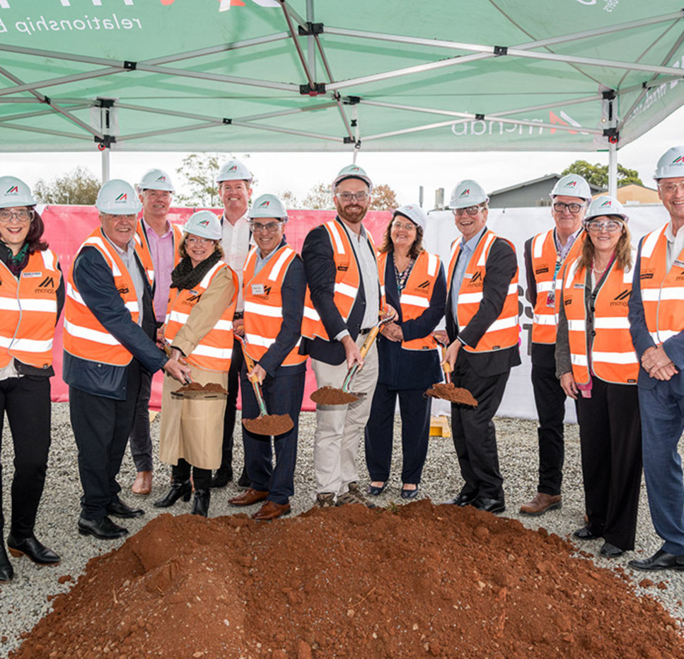 Group of people wearing safety vests and hard hats at a groundbreaking ceremony, smiling and holding shovels with soil in front of a construction site.