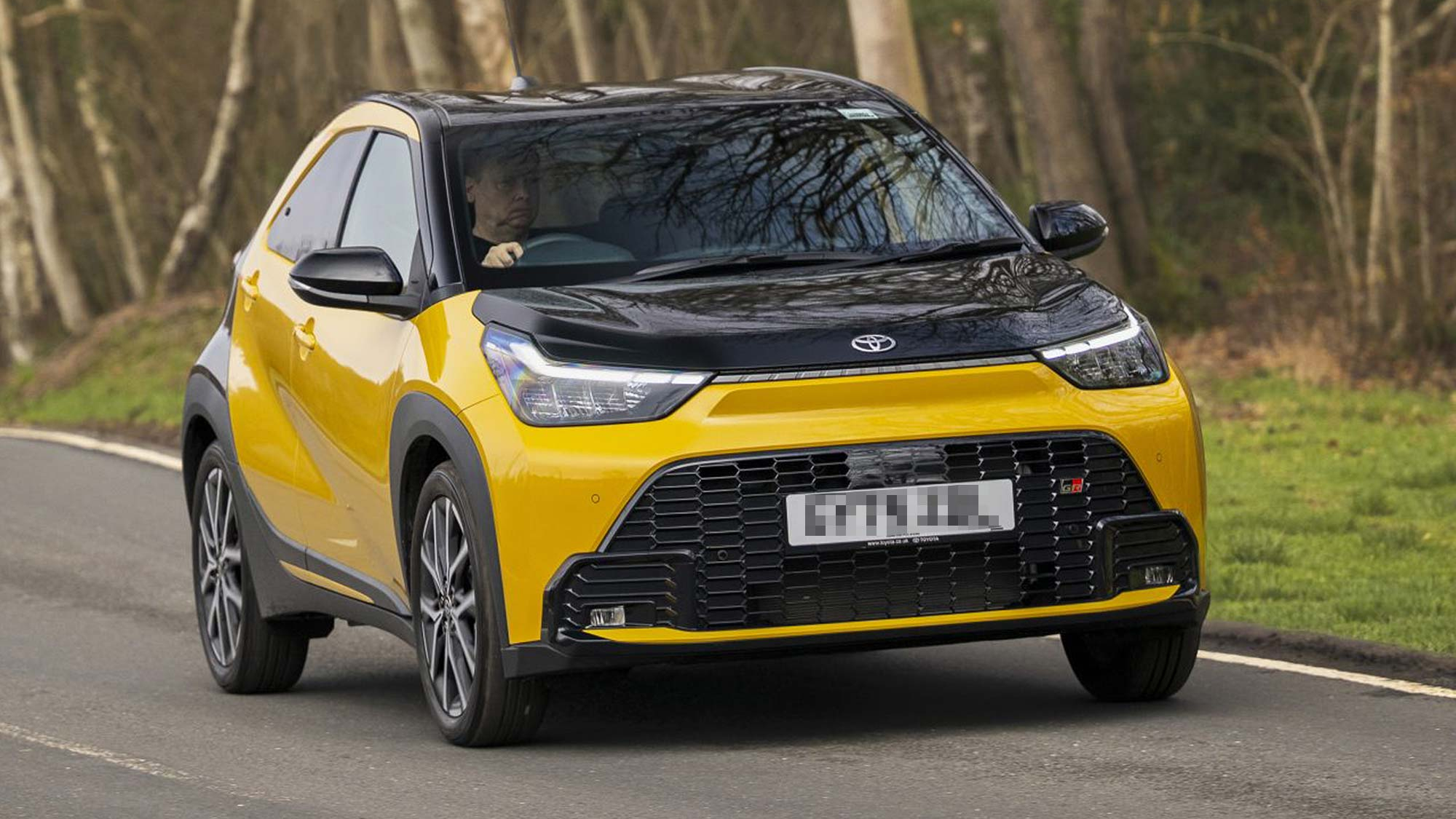 Yellow Toyota Aygo X city car driving on a road through woodland, shown from the front with a black roof.