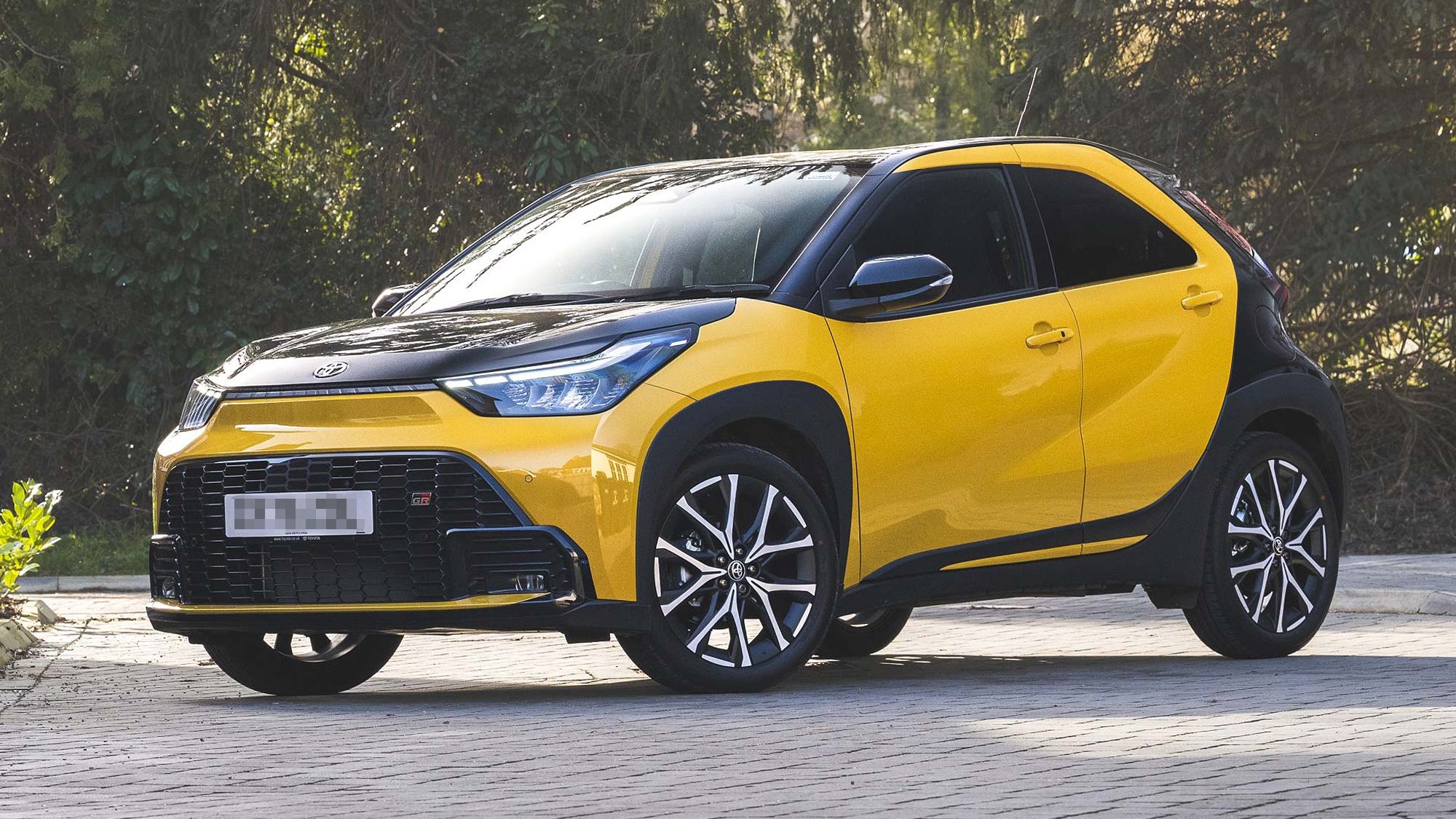 A yellow Toyota Aygo X parked on a driveway with trees in the background, front three quarter view, available on the Motability Scheme.
