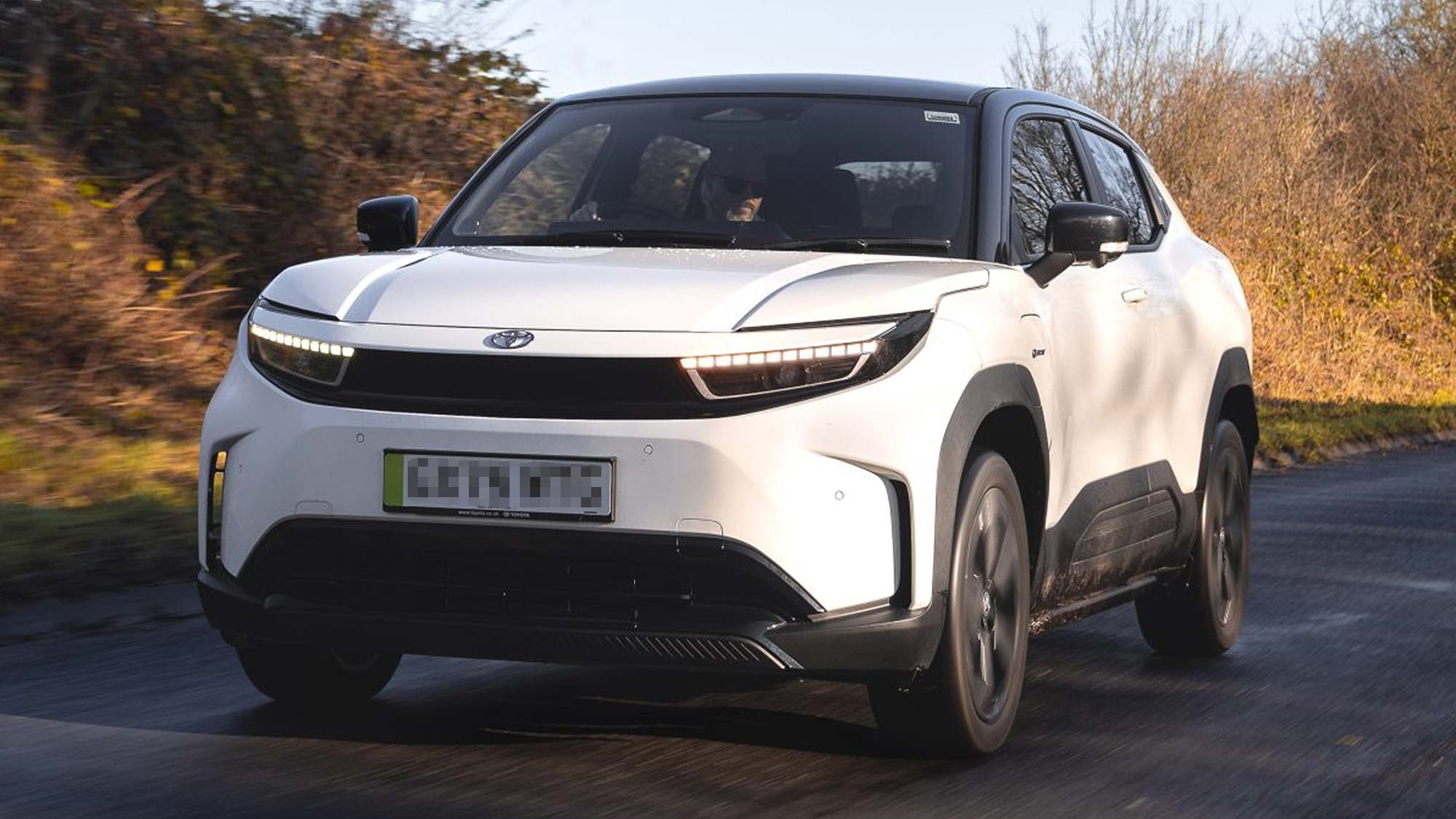 White Toyota Urban Cruiser driving along a rural road with trees and hedges, available on the Motability Scheme with no Advance Payment.