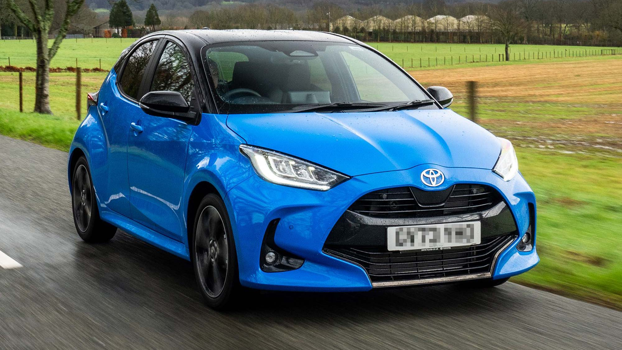 A blue Toyota Yaris hybrid hatchback is driving along a country road with fields and trees in the background.