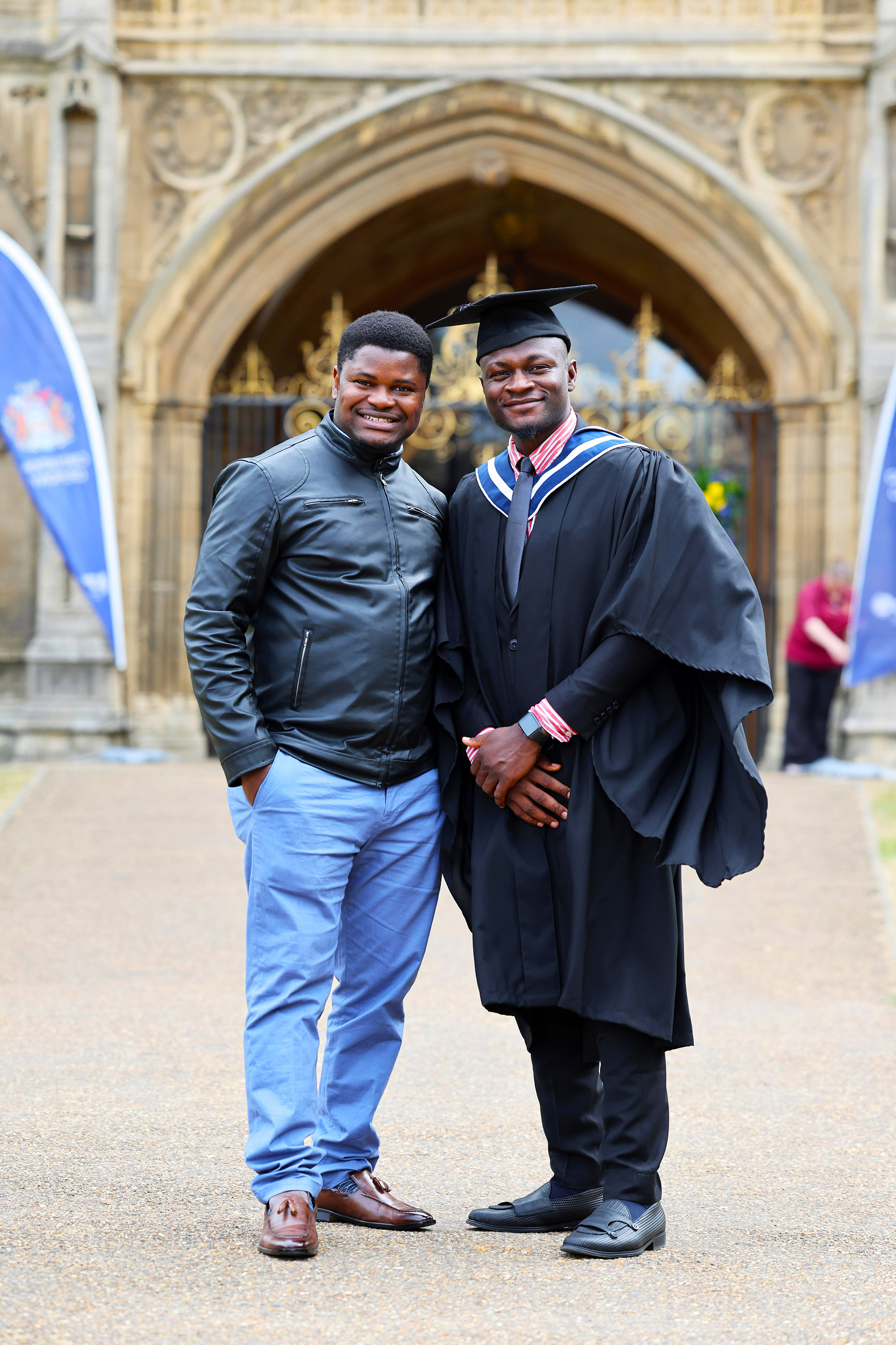 Two men posing outside a stone archway, one in a graduate gown