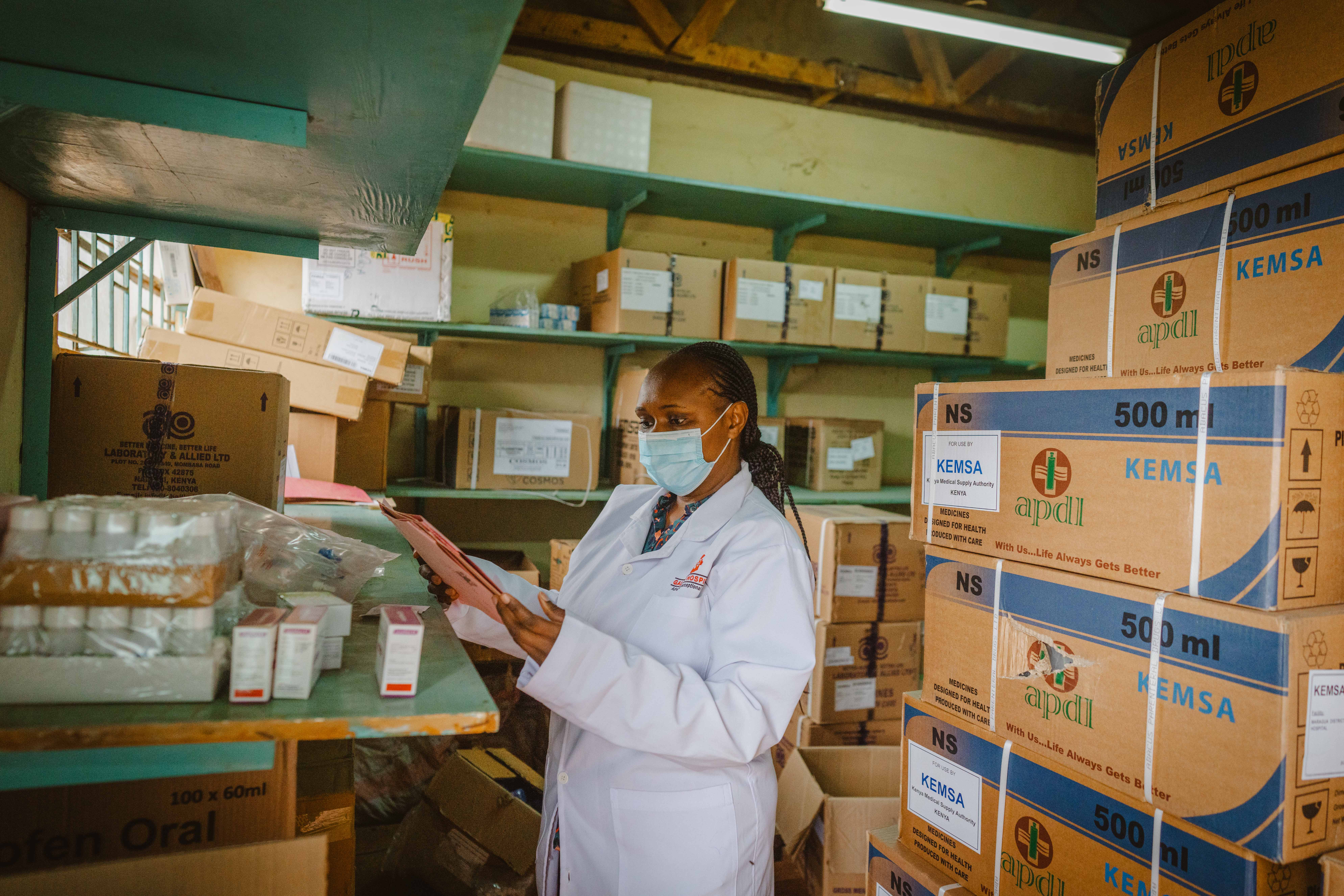 Woman in white doctors coat wearing a mask, standing next to storage boxes, taking items from a shelf