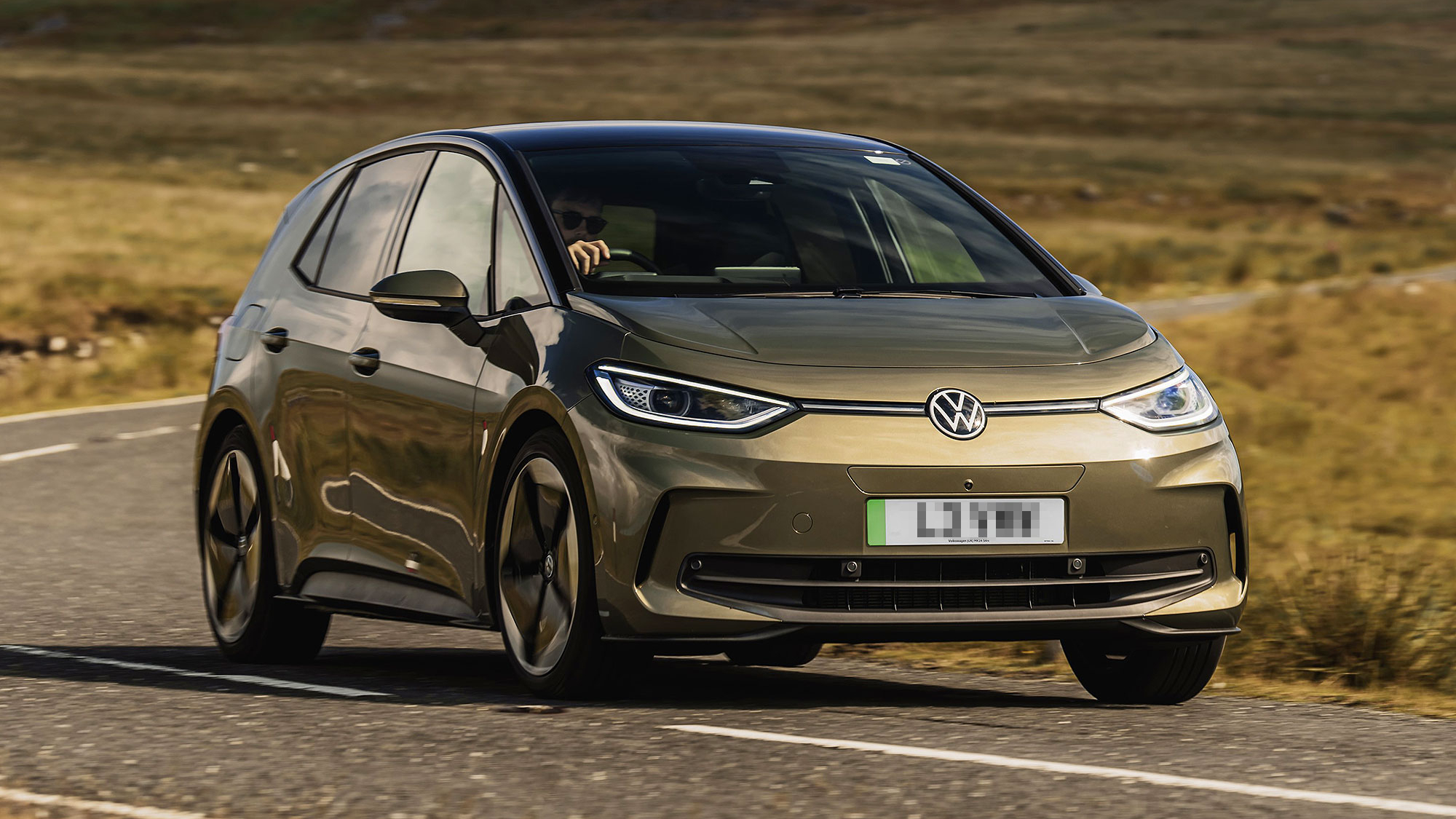 A bronze Volkswagen ID.3 electric hatchback driving on a rural road, with smooth aerodynamic styling and slim headlights.