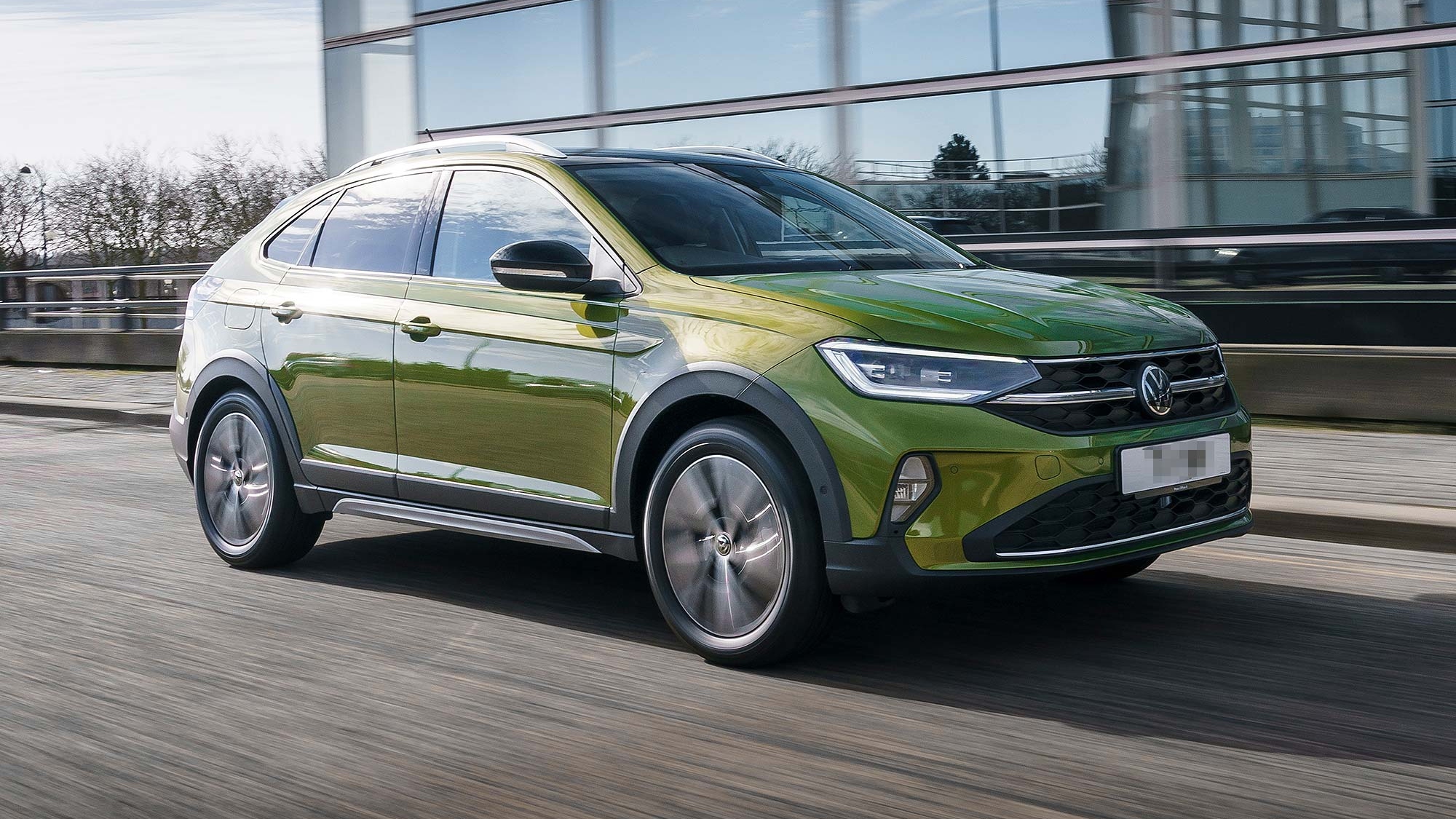Green Volkswagen Taigo SUV driving on a city road beside a modern glass building, available on the Motability Scheme with no Advance Payment.