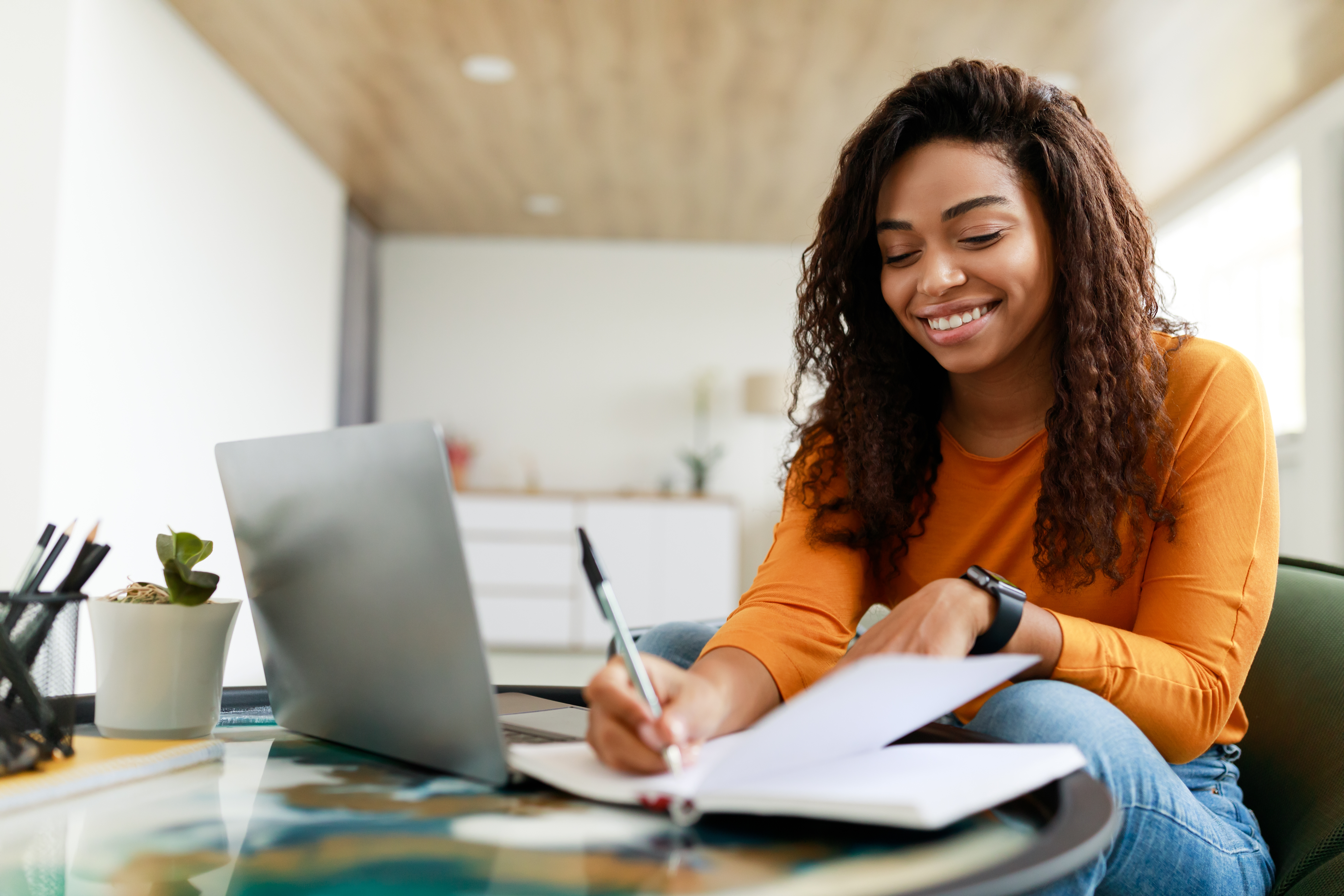 A young woman with curly dark hair wearing a long-sleeved orange top sits at a desk, smiling as she writes in an open notebook with a pen while working on a laptop.