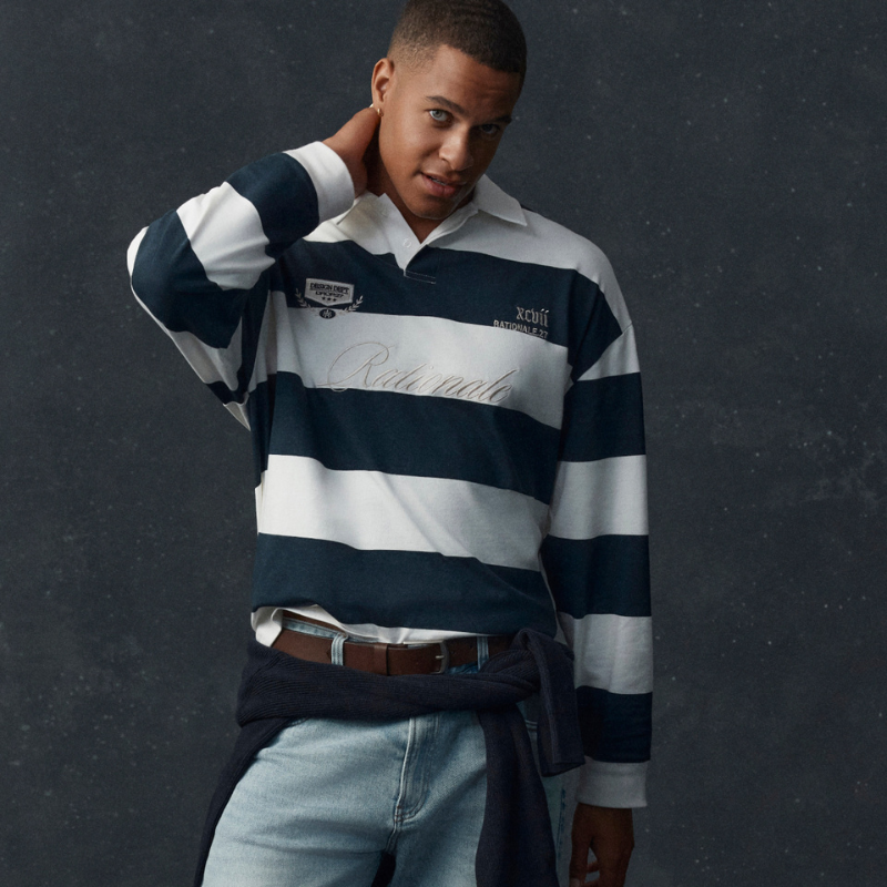 Model wearing a navy and white striped long sleeve rugby polo with light wash jeans, a brown belt and a navy knit tied around the waist.