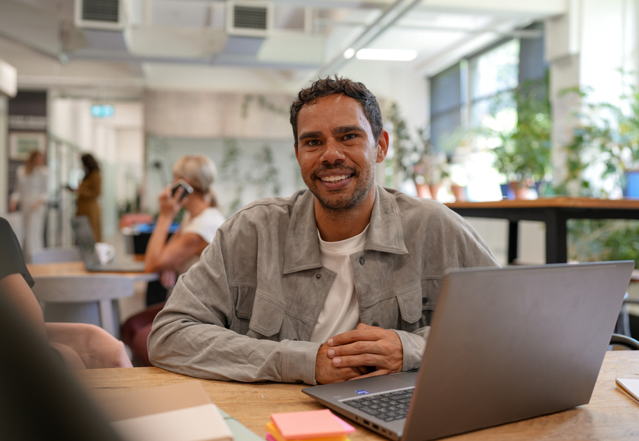 A man sits at a desk with a laptop, smiling at the camera in a bright, modern office. In the background, a woman is on the phone and there are green plants and large windows.