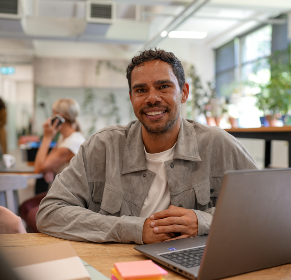 A man sits at a desk with a laptop, smiling at the camera in a bright, modern office. In the background, a woman is on the phone and there are green plants and large windows.