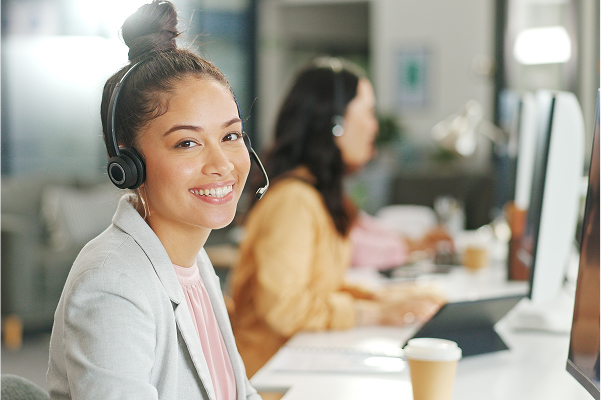 A smiling female customer service agent wearing a headset looks directly at the camera while seated at her desk in a modern office, with a colleague working in the background.