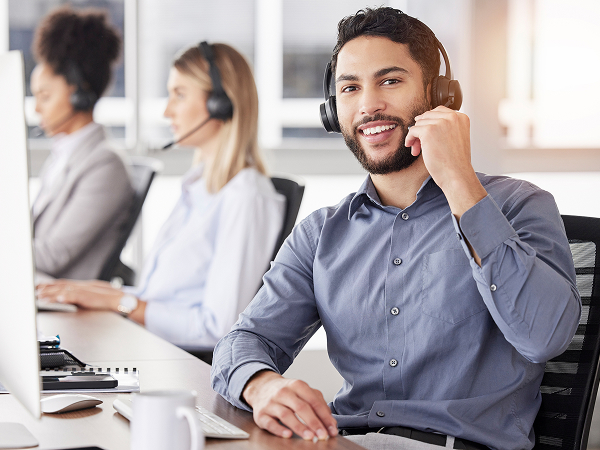 A smiling male customer service agent wearing a headset and a grey button-down shirt looks at the camera while working at a desk in a busy call center office, with two other agents in the background.