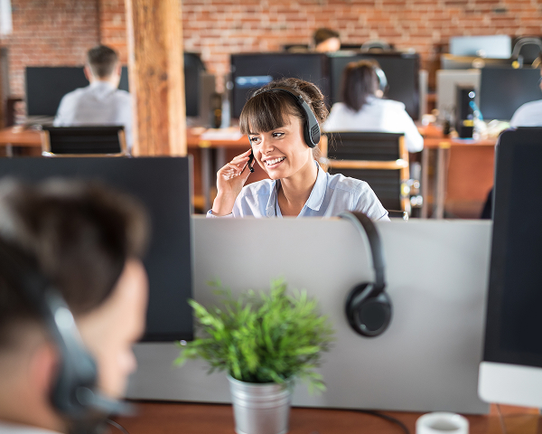A smiling female customer service agent wearing a headset looks to the side while talking, seated at a desk in an open-plan office with exposed brick walls. Computer monitors and a small plant are visible in the foreground.