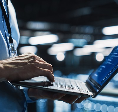 Close-up of a person's hands typing on a laptop keyboard in a dimly lit, high-tech server room or data center, with blue lights blurring in the background.