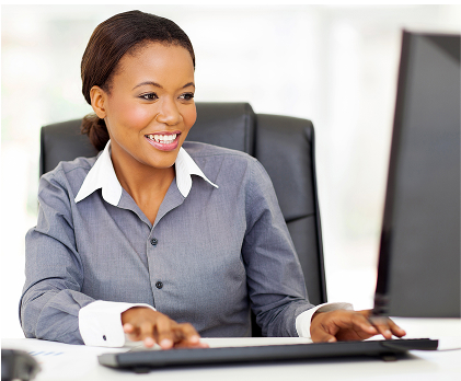 A smiling woman in a grey business shirt is seated at a desk, typing on a keyboard while looking at a computer monitor off-screen.