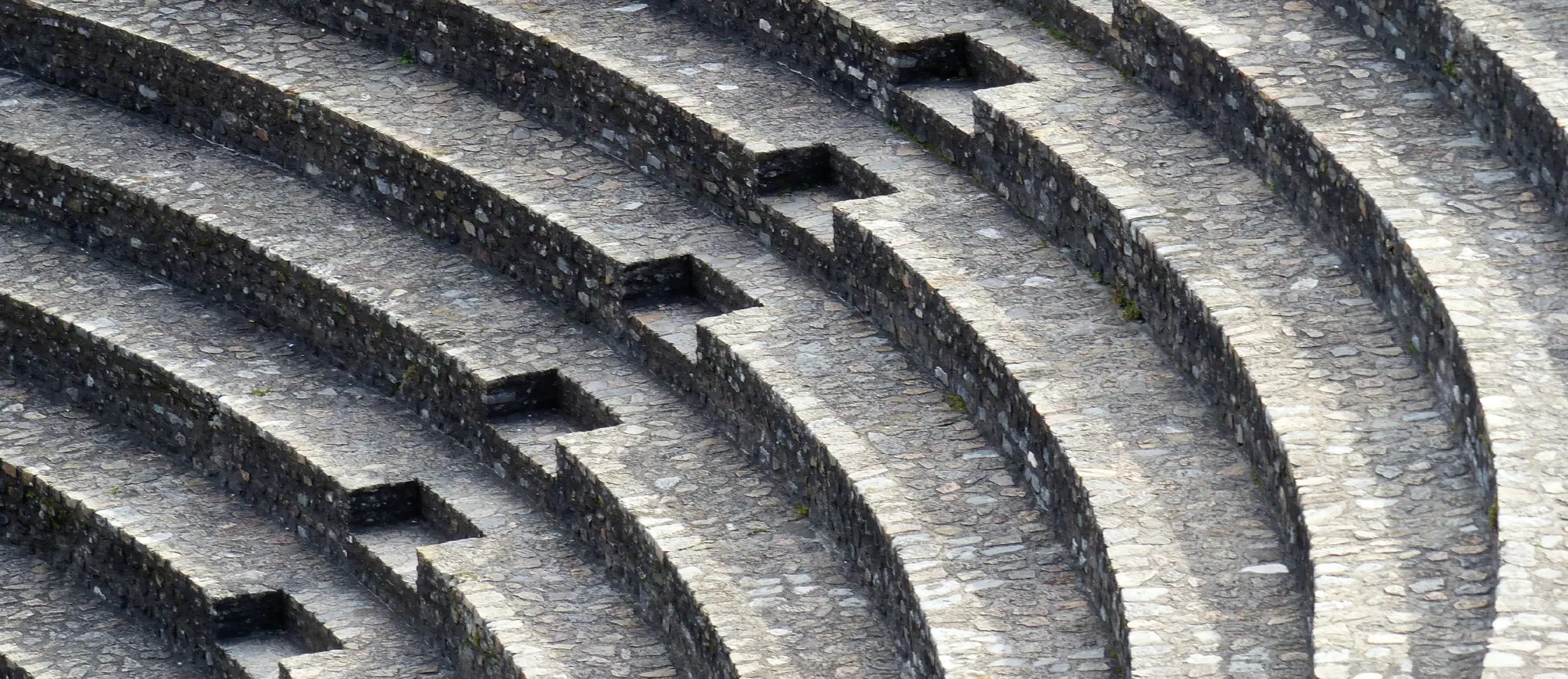 An abstract, high-angle view of semicircular, terraced steps made of rough-cut grey and beige stones. The steps curve outward and upward from the bottom left to the top right. A small, straight flight of stairs cuts across the curve at multiple points, connecting the terraced levels in a repeating, staggered pattern, creating a strong sense of texture and depth.