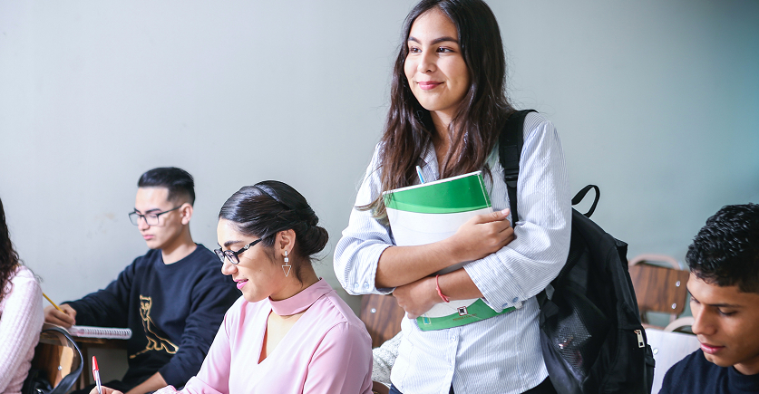 A student, standing and smiling, holds a green and white folder and books while wearing a backpack in a classroom. Other students are seated and writing at desks in the foreground.